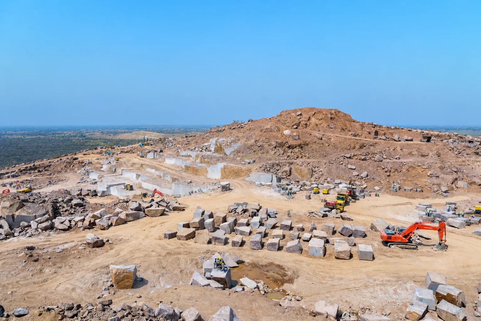 Aerial view of a quarry hill with cut stone blocks and an excavator under a blue sky